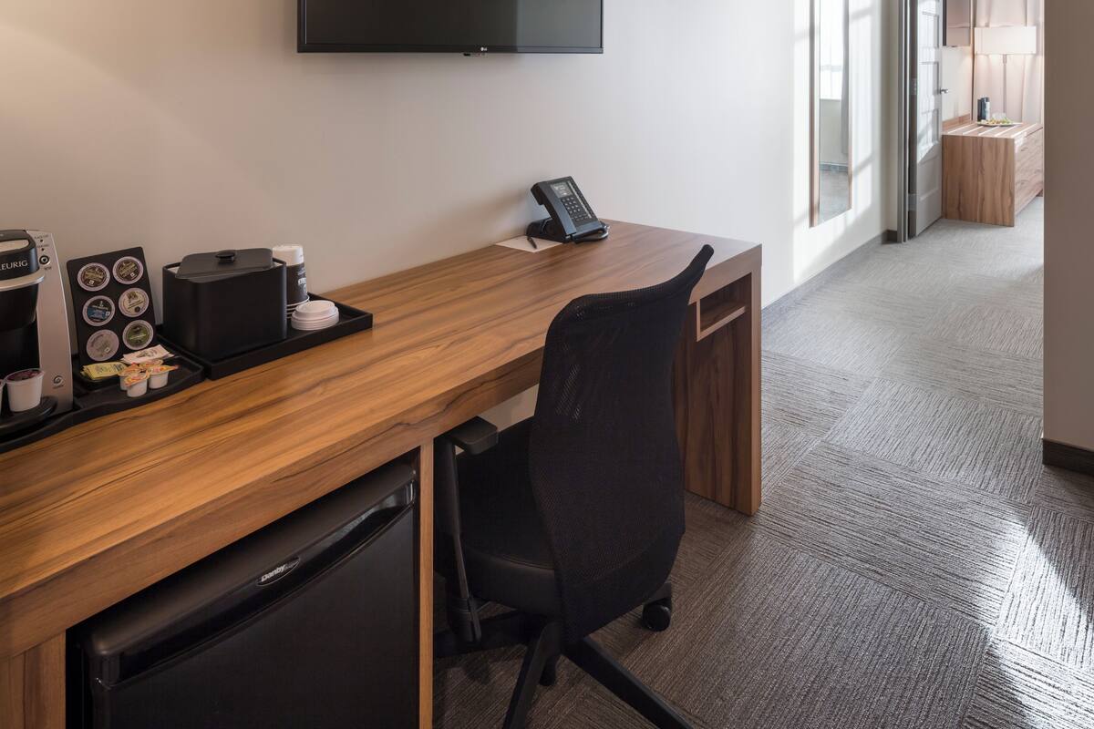 Habitación de hotel con un escritorio de madera, una silla de oficina negra, una cafetera y un televisor de pantalla plana montado en la pared.