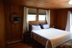 Habitació amb panells de fusta, un llit, una tauleta de nit, un mirall i una finestra amb cortines.