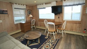 A cabin interior with a dining table, chairs, a sofa, a TV mounted on the wall, and a window overlooking a body of water. - Belknap Point Inn (Gilford)