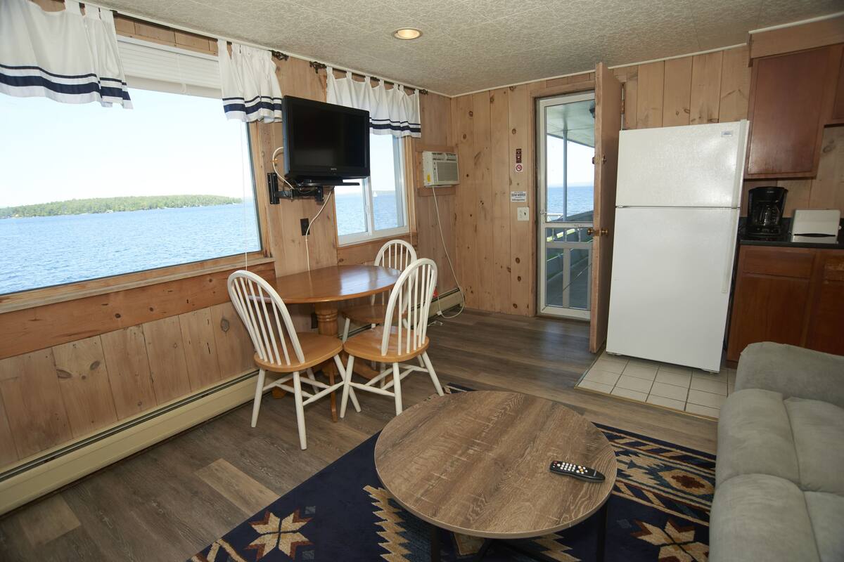 A compact cabin with a dining area, kitchen, and a large window offering a view of the water.