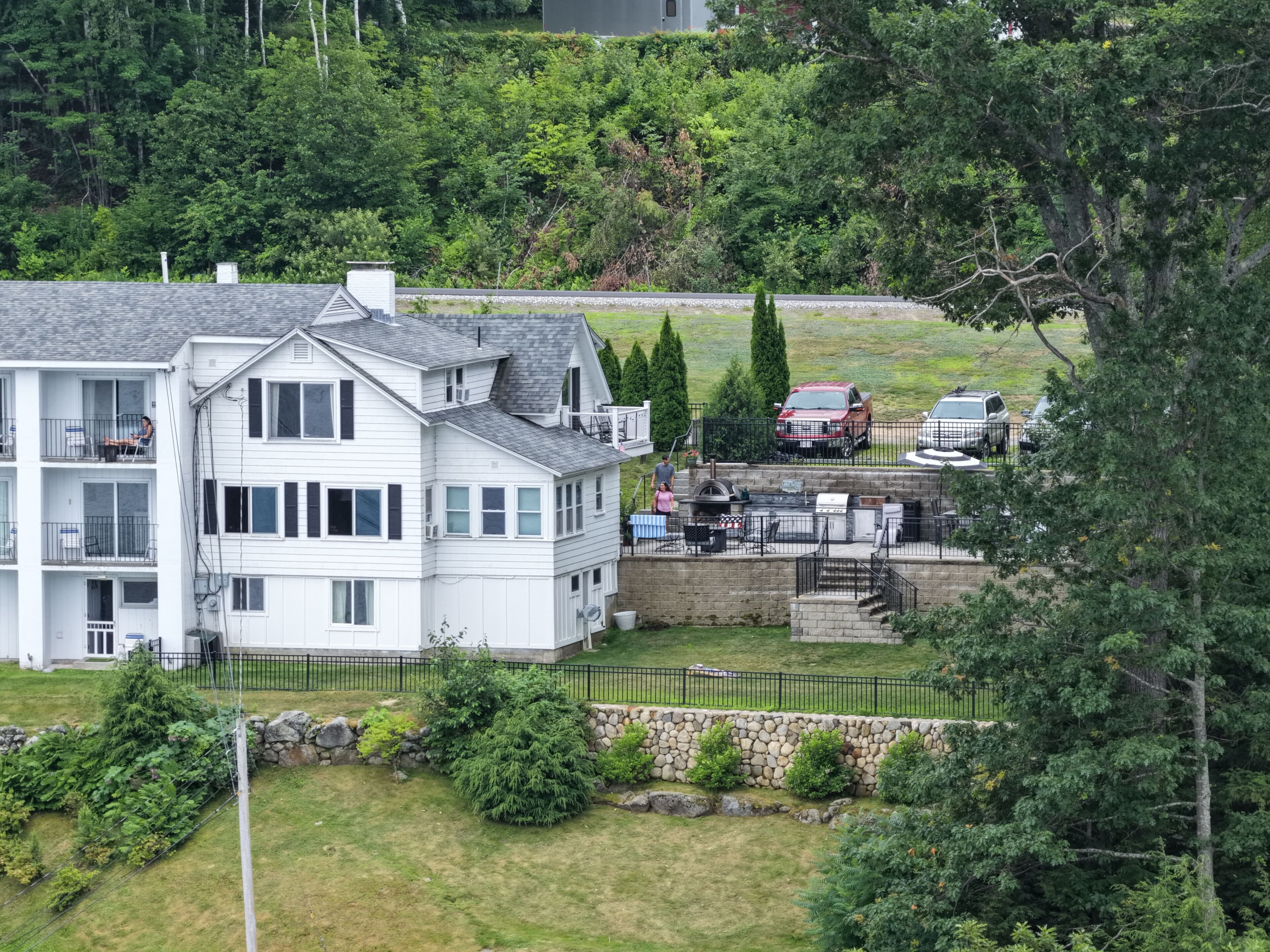 A multi-story white building with a balcony, a stone retaining wall, and a fenced yard with parked cars.