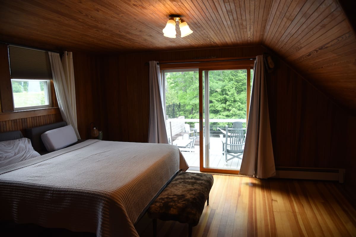 A bedroom with a large bed, a bench, and a sliding glass door leading to an outdoor area with chairs.
