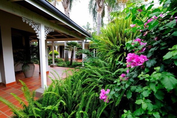 A covered outdoor area with a tiled floor, surrounded by lush greenery and flowering plants.