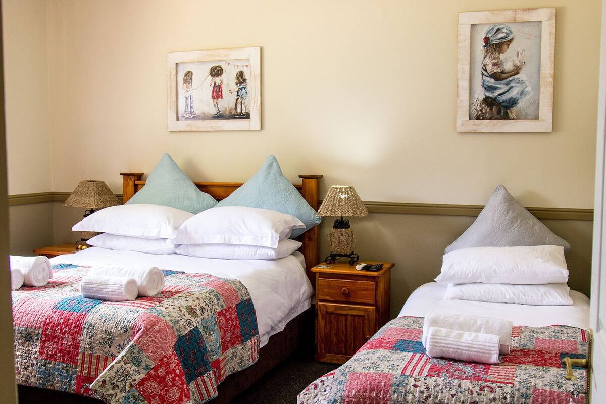 A hotel room with two beds, each with a quilted bedspread, pillows, and a nightstand with a lamp. There are framed pictures on the wall above the beds.