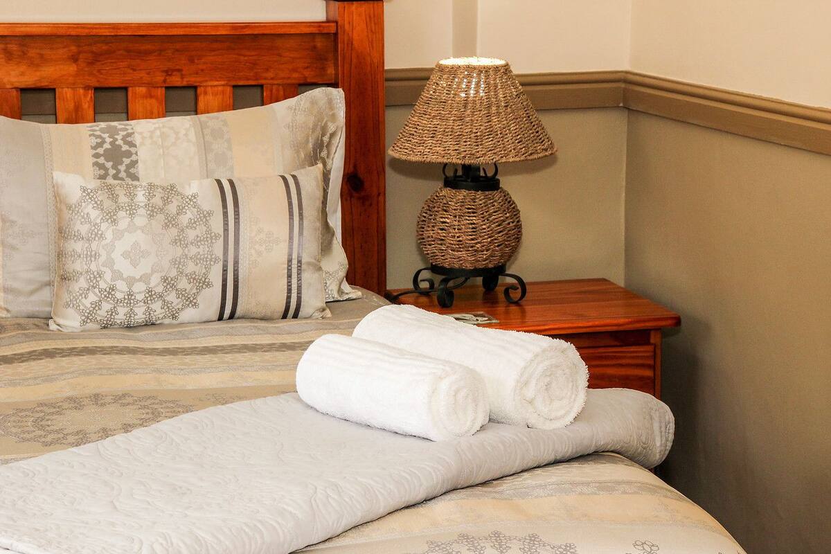 A neatly made bed with patterned pillows, a rolled-up towel, and a woven table lamp on a bedside table.
