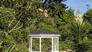 Ein mit Gitterwerk versehenes Gazebo, umgeben von üppigem Grün und blühenden Bäumen.