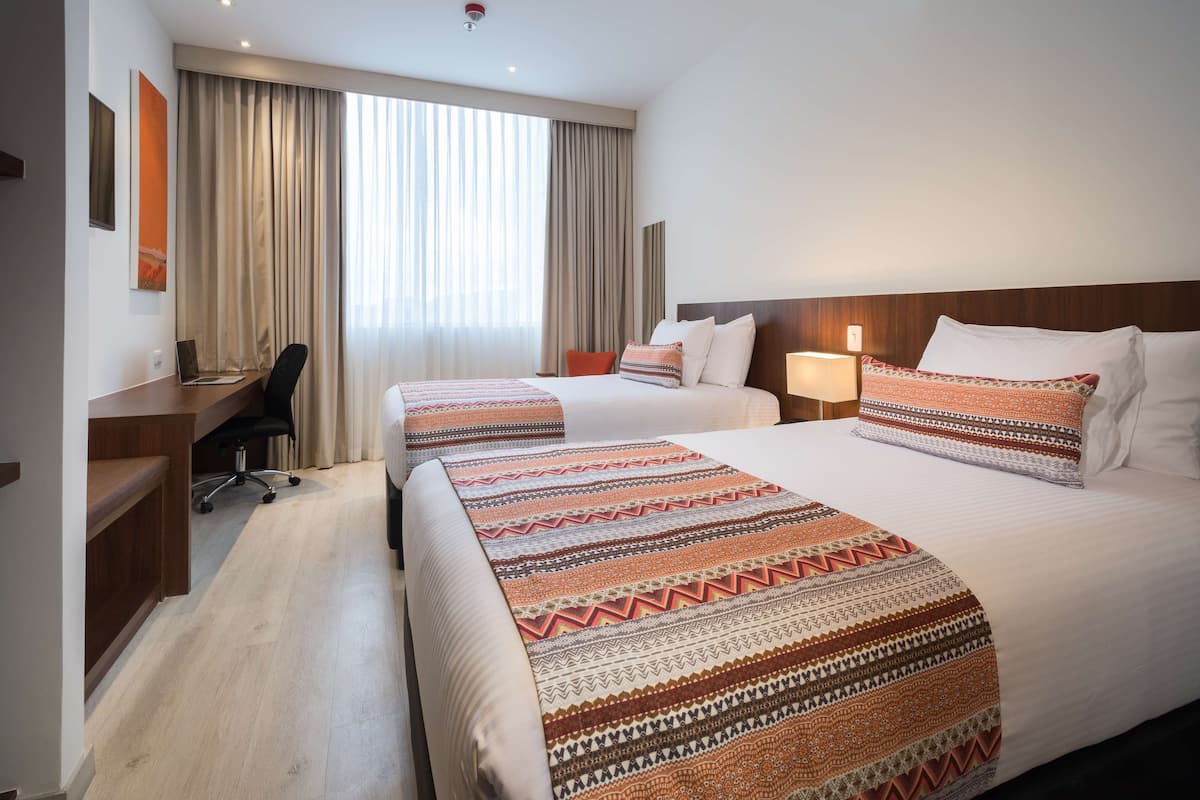 Quarto de hotel com duas camas, uma escrivaninha e uma cadeira.