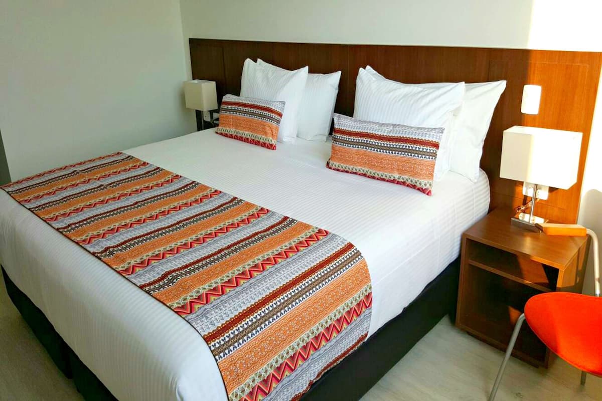 Quarto de hotel com uma cama grande, roupa de cama branca, manta estampada e mesa de cabeceira de madeira com uma luminária.