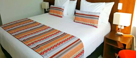Quarto de hotel com uma cama grande, roupa de cama branca, manta estampada e mesa de cabeceira de madeira com uma luminária.
