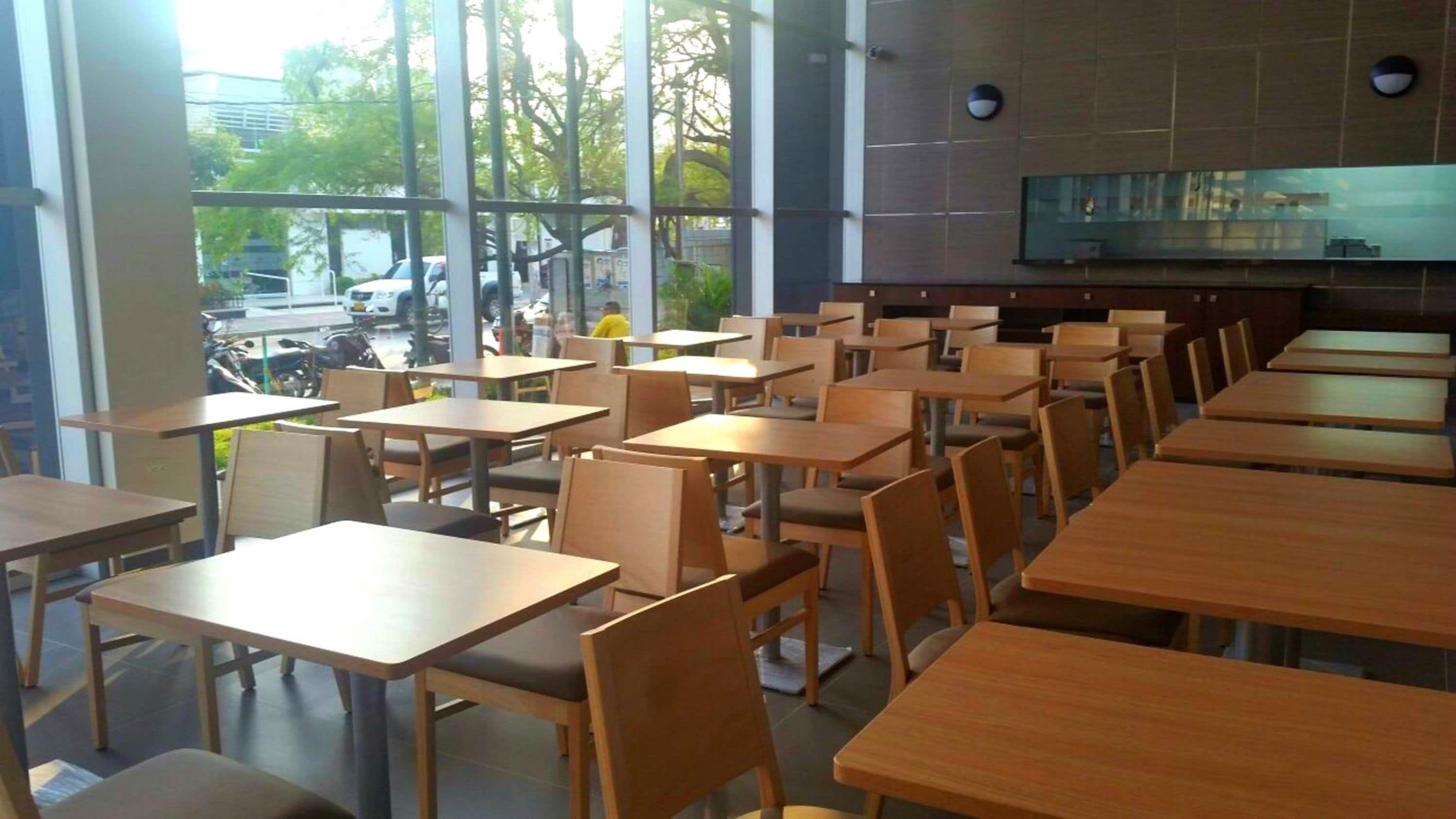 Een modern restaurant met houten tafels en stoelen, grote ramen met uitzicht op de straat en geparkeerde auto's.