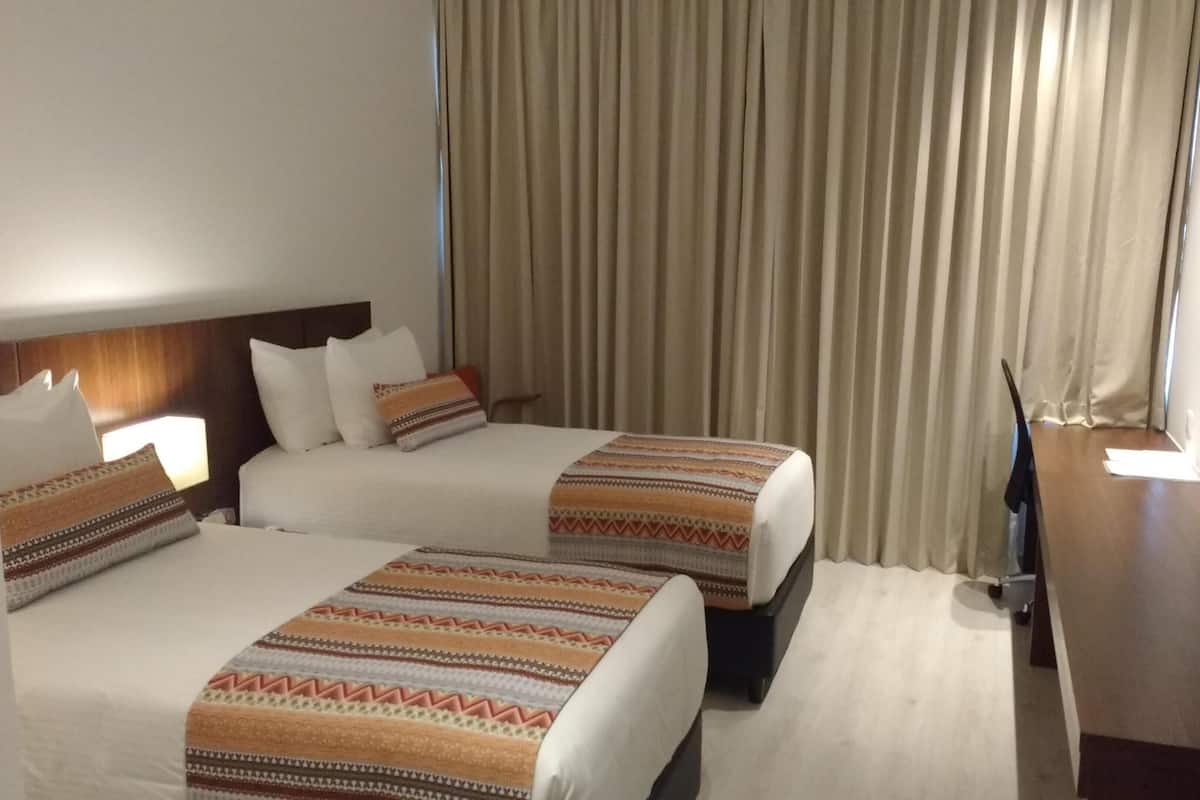 Quarto de hotel com duas camas, uma escrivaninha, uma televisão e uma janela grande com cortinas.