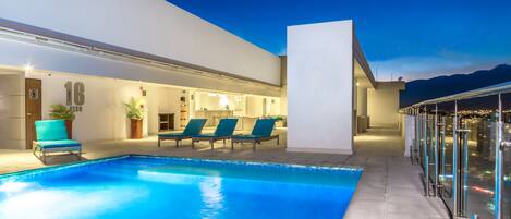 A modern hotel with a swimming pool, lounge chairs, and a glass-enclosed balcony.