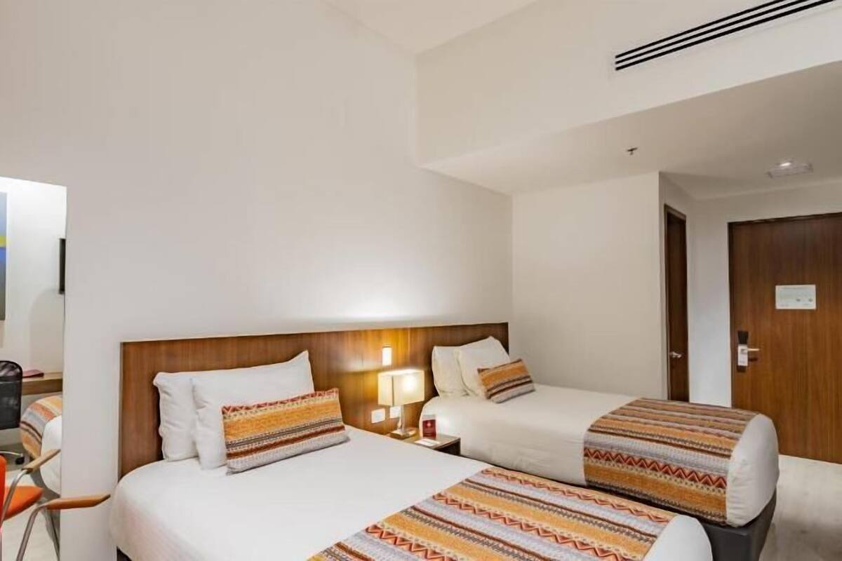 Quarto de hotel com duas camas, uma escrivaninha, uma cadeira e uma porta.