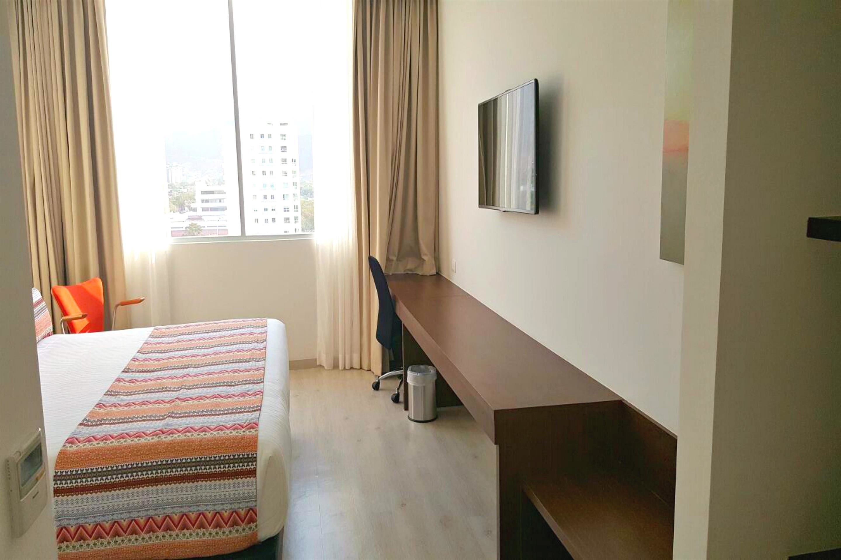A hotel room with a bed, a desk, a chair, a TV, and a window with curtains.