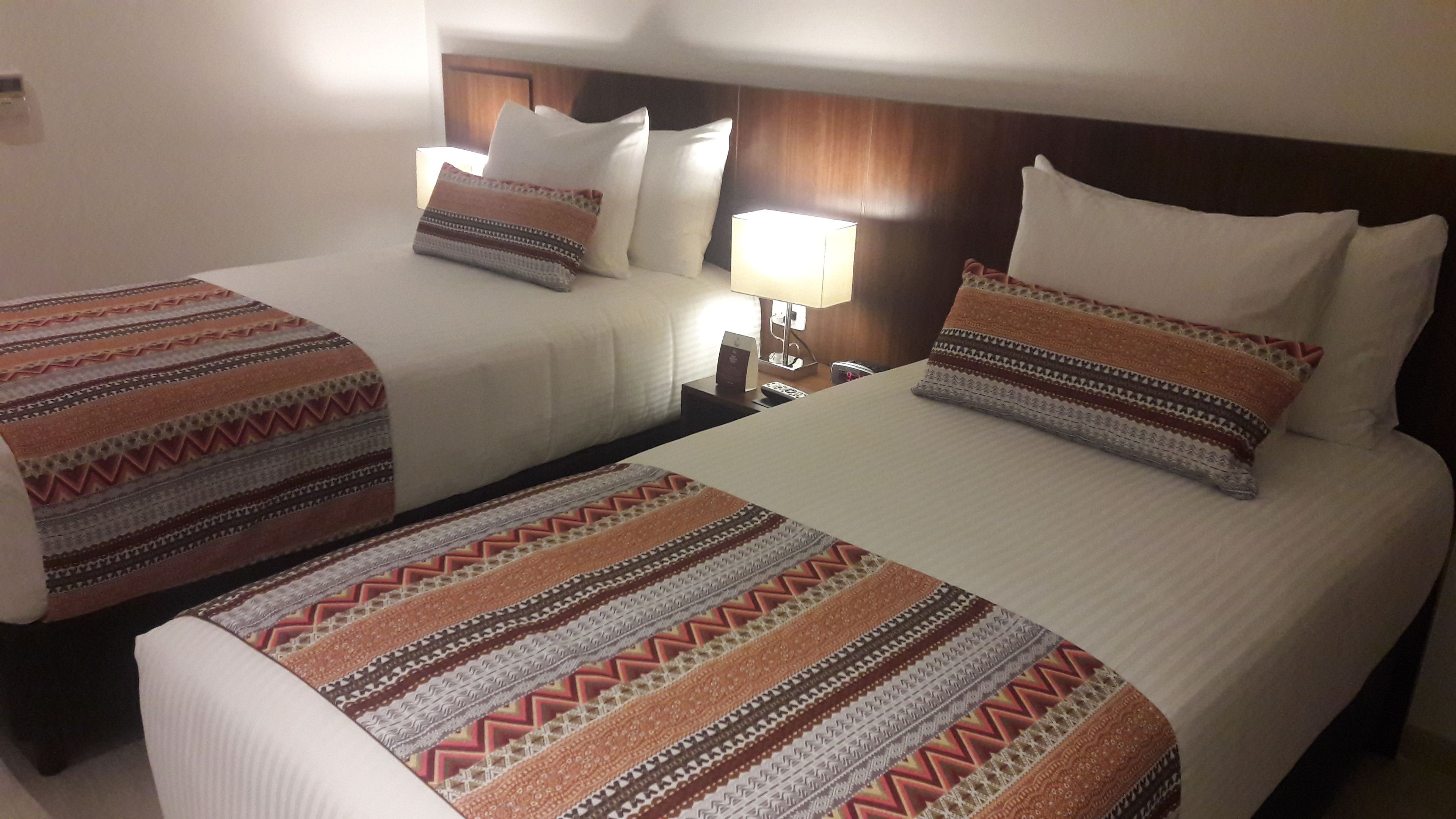 A hotel room with two beds, each with patterned blankets and pillows. There is a nightstand with a lamp between the beds.