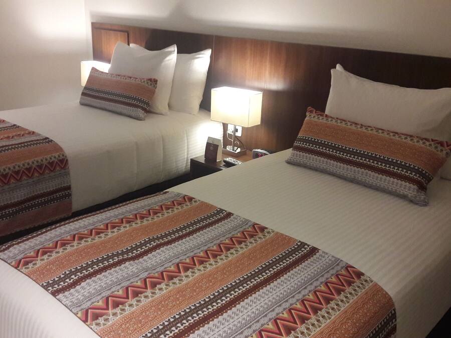 Habitación de hotel con dos camas, cada una con mantas y almohadas estampadas. Hay una mesita de noche con una lámpara entre las camas.