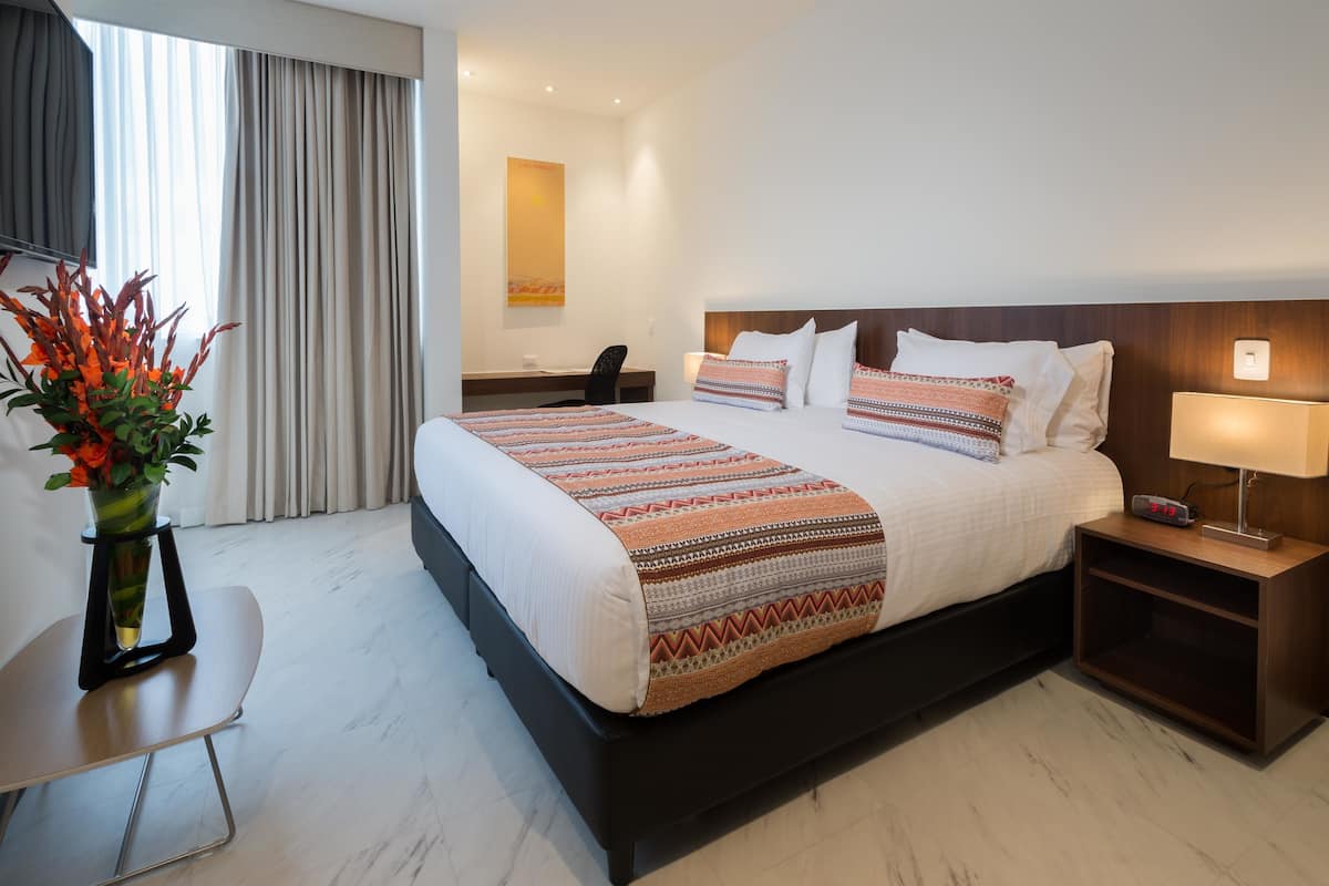 Quarto de hotel com cama grande, uma escrivaninha, uma cadeira, mesa de cabeceira e um vaso com flores.