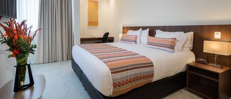 A hotel room with a large bed, a desk, a chair, a nightstand, and a vase with flowers.