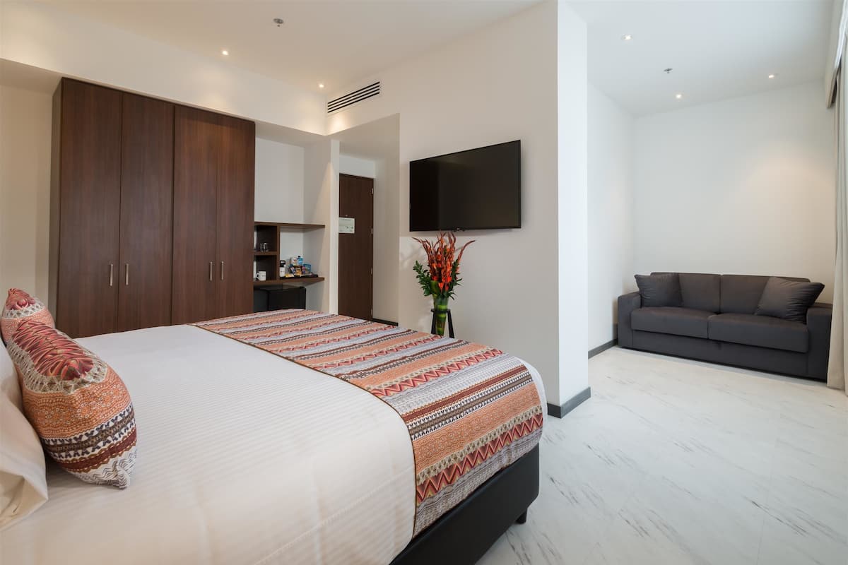Um quarto moderno com uma cama grande, um guarda-roupa de madeira escura, um sofá cinzento e uma televisão de tela plana fixada na parede.