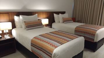 A hotel room with two beds, each with patterned bedspreads and white pillows. There are bedside tables with lamps and a clock.