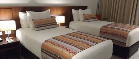 Habitación de hotel con dos camas, cada una con cubrecamas estampados y almohadas blancas. Hay mesitas de noche con lámparas y un reloj.