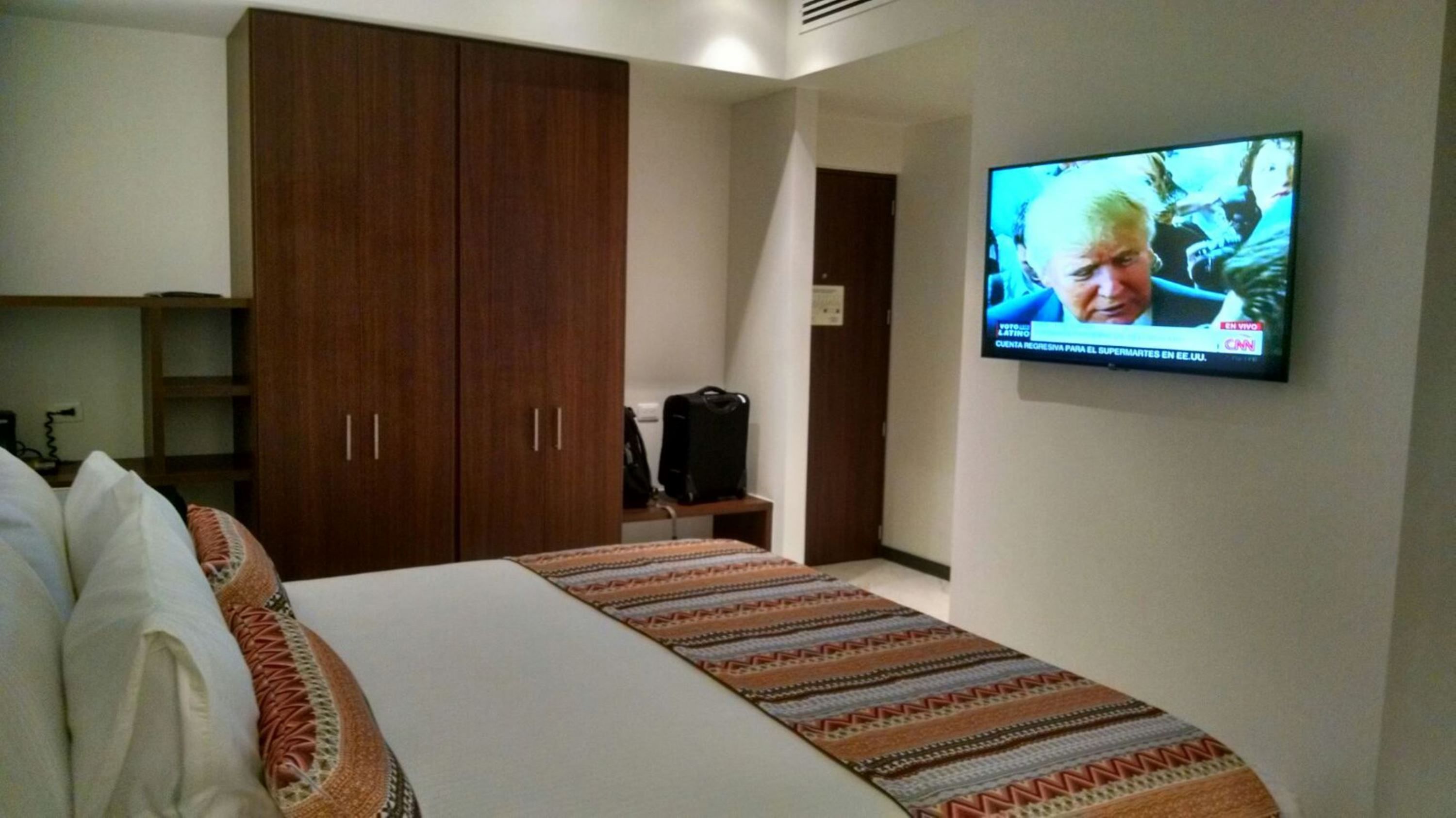 Una habitación de hotel con una cama, un televisor en la pared, un armario y una maleta sobre una mesita.