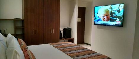Ett hotellrum med en säng, en tv som är monterad på väggen, en garderob och en resväska på ett litet bord.