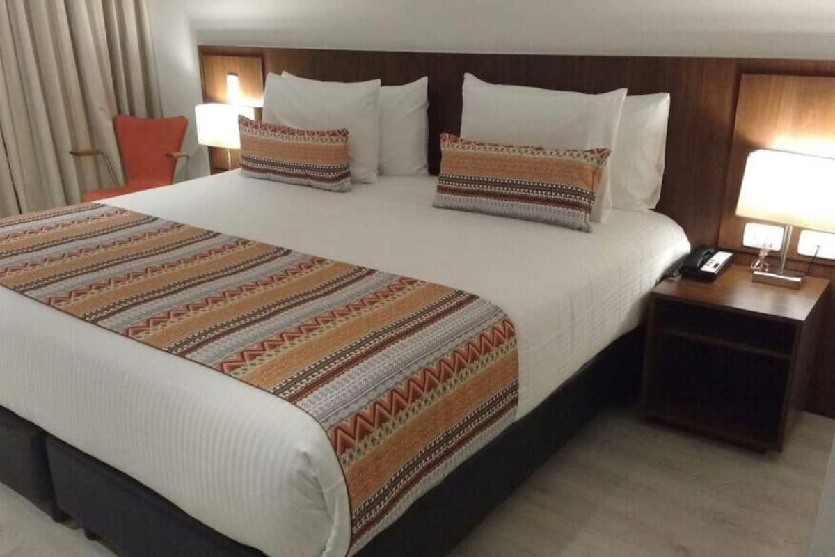 Quarto de hotel com uma cama grande, duas mesas de cabeceira, uma cadeira e uma janela com cortinas.