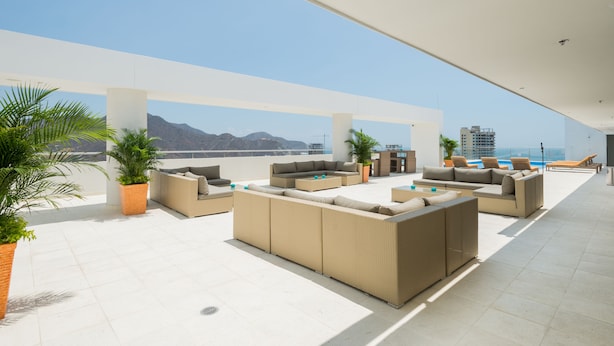A rooftop lounge area with outdoor furniture, potted plants, and a view of the ocean and mountains.