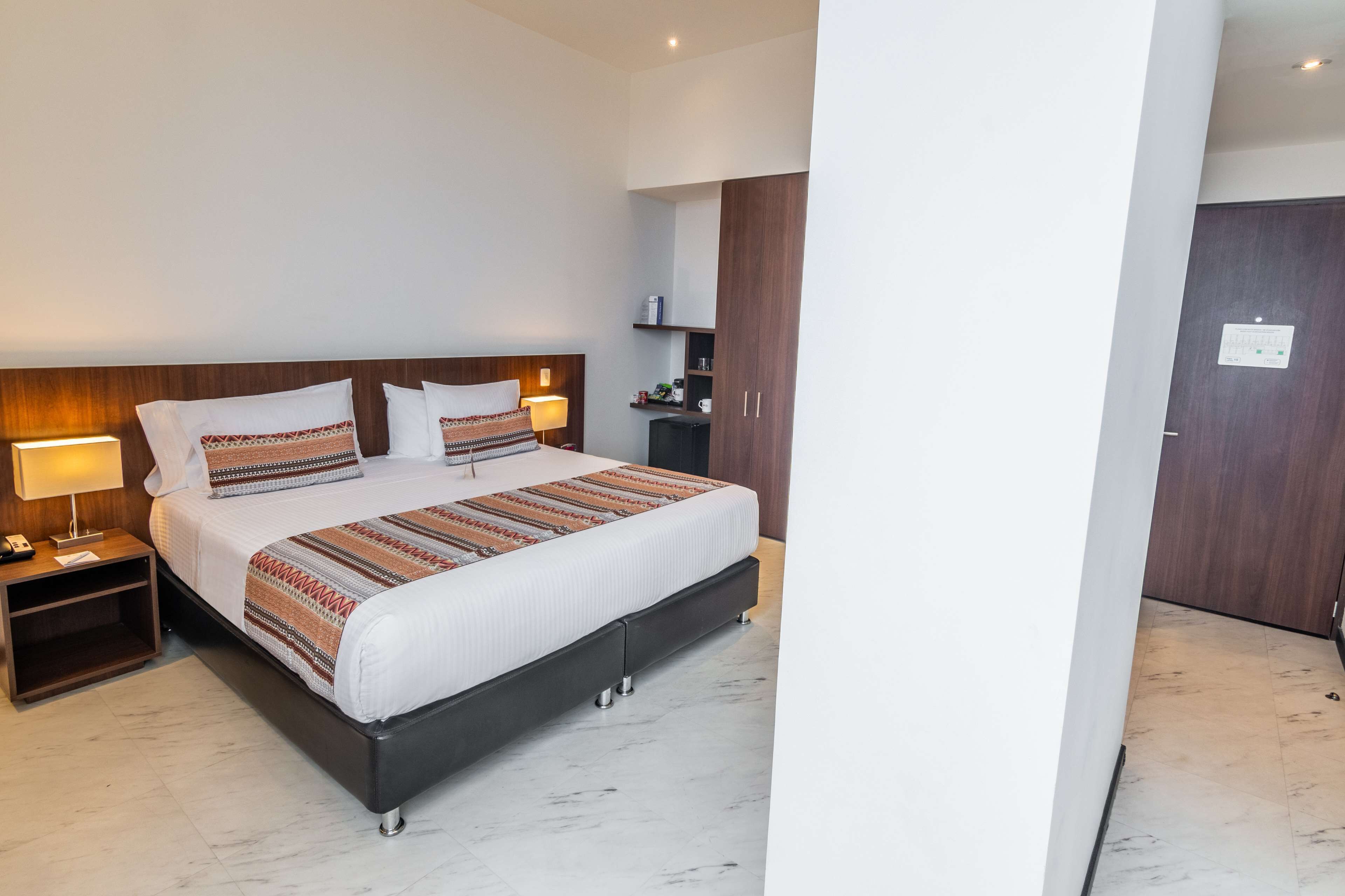 Une chambre d’hôtel avec un grand lit, une tête de lit en bois, des tables de chevet avec des lampes, une armoire et une petite étagère avec des articles de toilette.