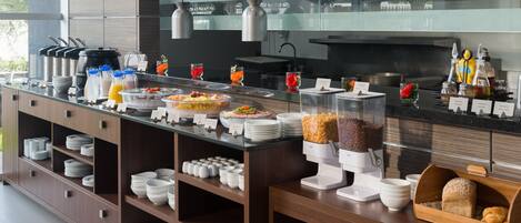 Una barra de buffet con diversos alimentos y bebidas, incluyendo frutas, cereales y refrescos, dispuesta sobre una encimera con una cocina moderna al fondo.