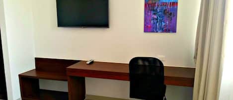 A room with a wooden desk, a black office chair, a flat-screen TV mounted on the wall, and a painting above the desk.