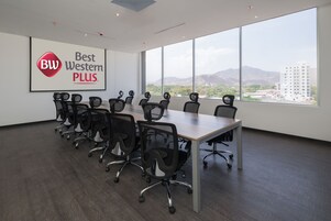 A conference room with a large table and multiple chairs, a large window with a view of mountains and buildings, and a sign on the wall reading "Best Western PLUS".