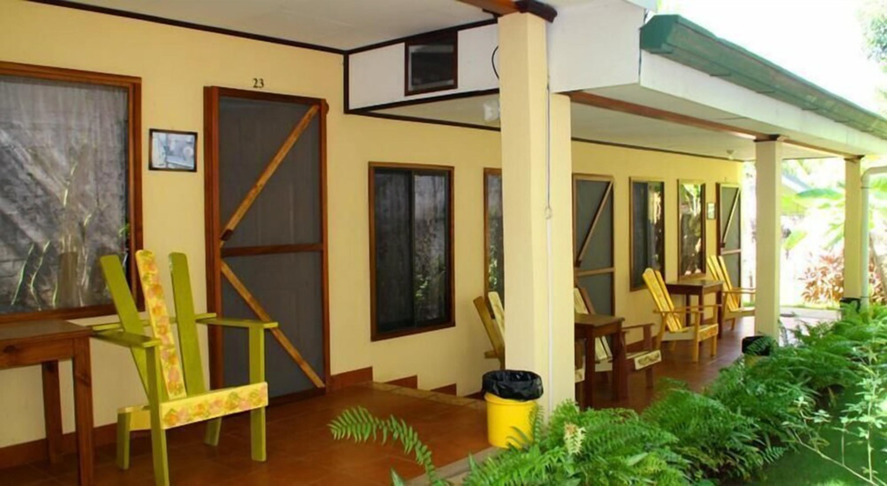 Un área de estar al aire libre techada, con sillas y mesas de madera, un cubo de basura y una puerta con el número 23.