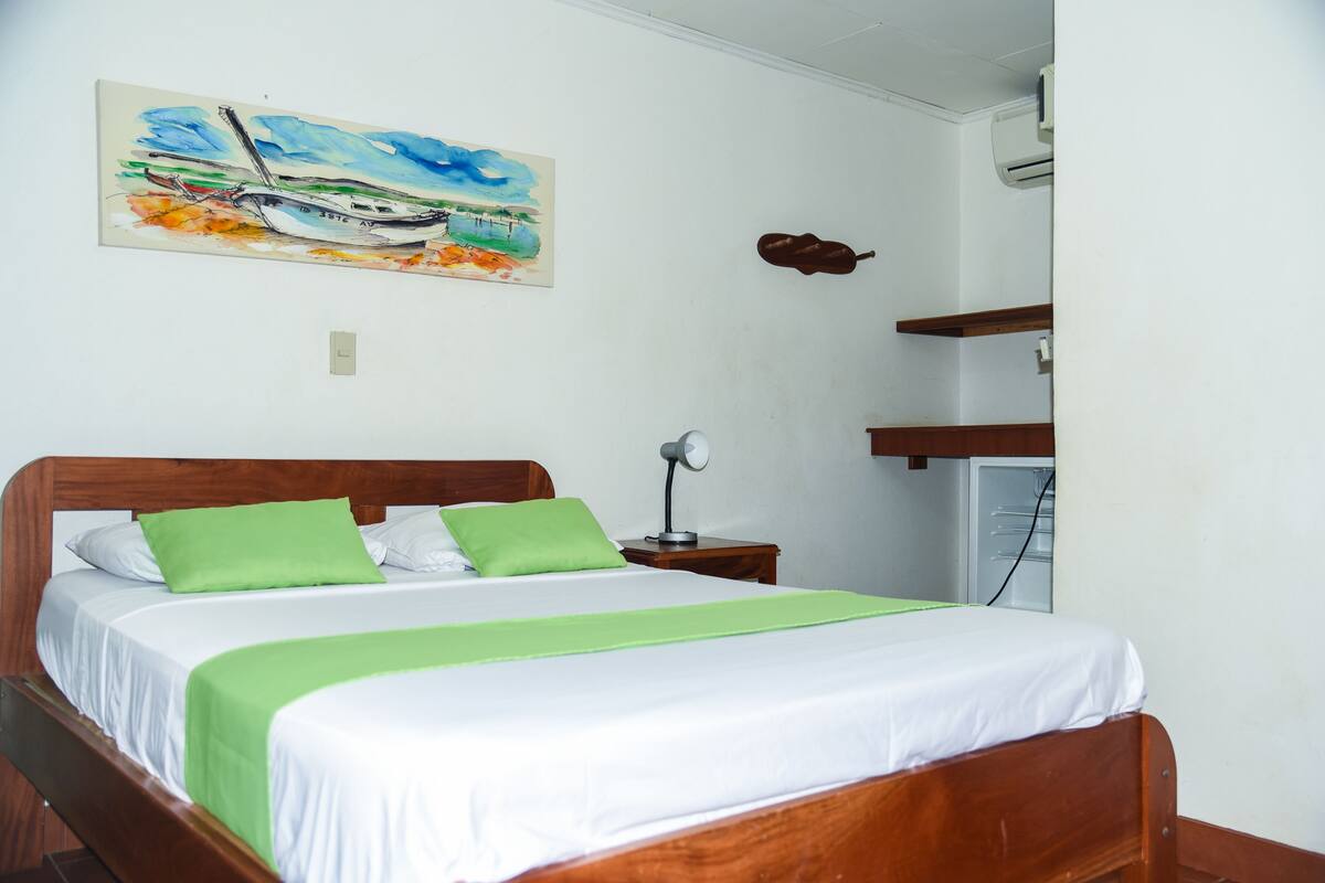 Ein Schlafzimmer mit einem Bett, grünen Kissen, einem Nachttisch mit Lampe, einem Bild eines Bootes und einer Gitarre an der Wand.