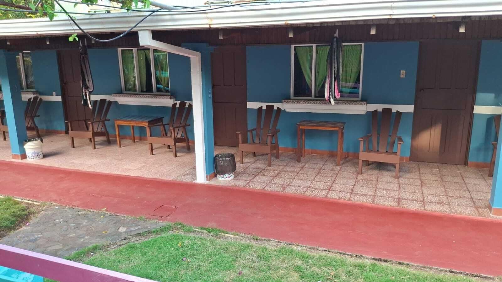 A covered outdoor seating area with wooden chairs and tables, a red pathway, and a building with blue walls and windows.