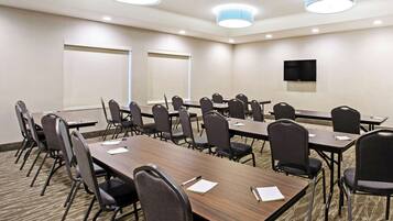 A conference room with multiple tables and chairs, a flat-screen TV mounted on the wall, and large windows with blinds.