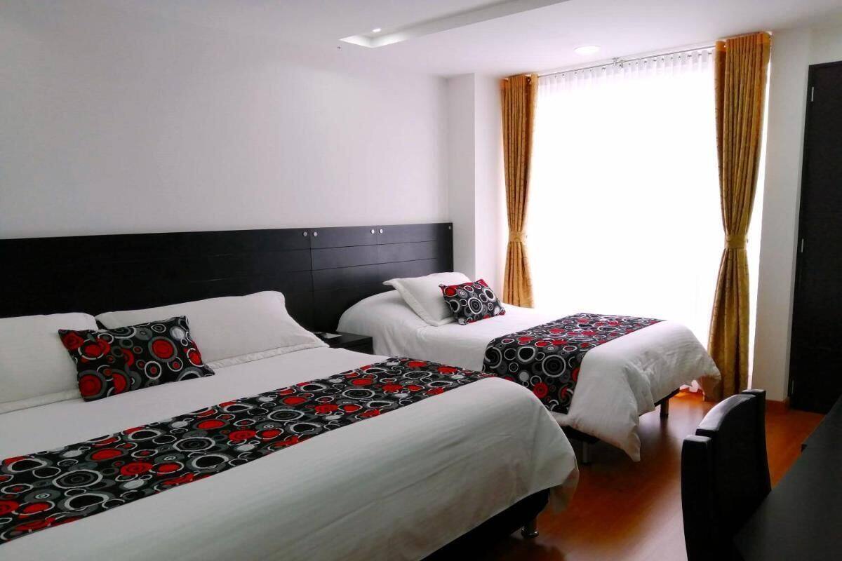 Habitación de hotel con dos camas, cada una con cubrecama blanco y mantas estampadas en negro y rojo. Tiene cabecera negra, ventana con cortinas y un escritorio con silla.
