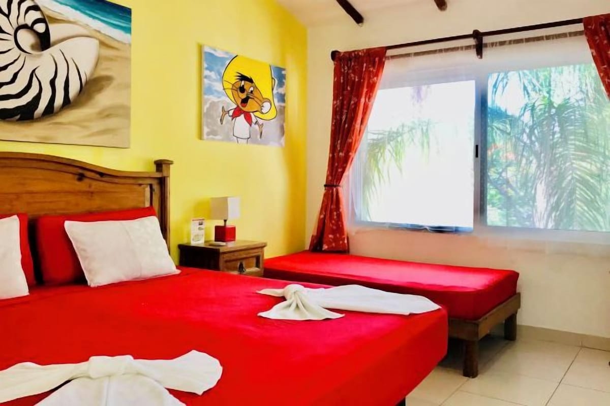 Habitación de hotel con dos camas, cabecera de madera, un cuadro de un personaje de dibujos animados, ventana con cortinas rojas y vistas a palmeras.