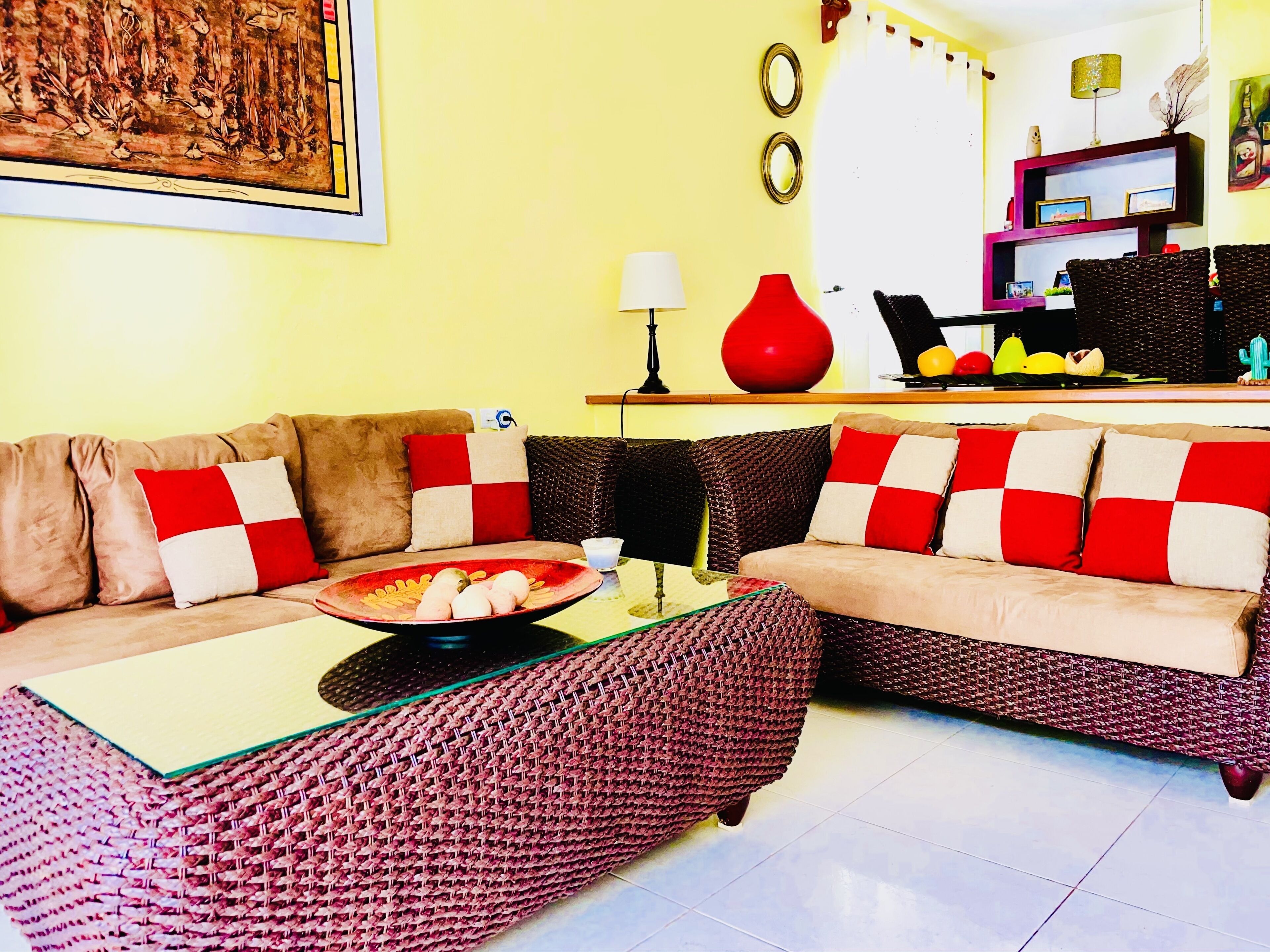 Una sala de estar con un conjunto de sofá, una mesa de centro y una zona de comedor con un jarrón rojo y una bandeja de frutas.