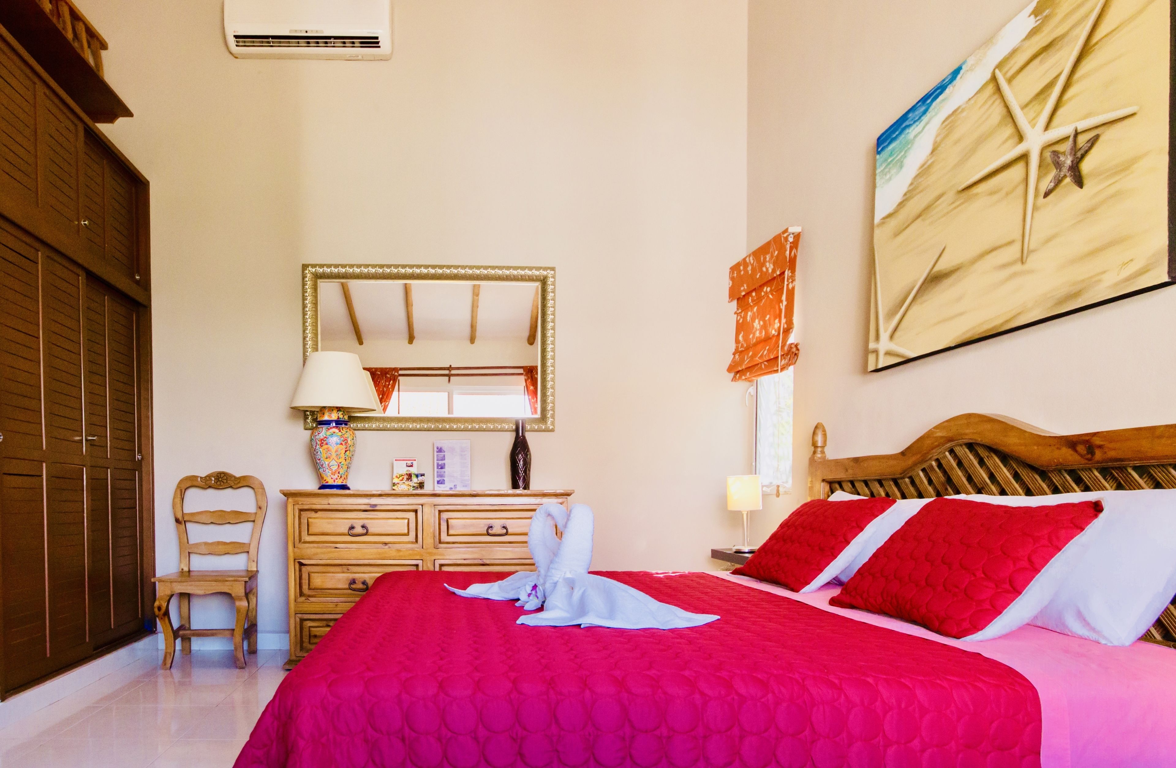 A hotel room with a large bed, wooden headboard, a dresser, a chair, a mirror, and a beach-themed painting.
