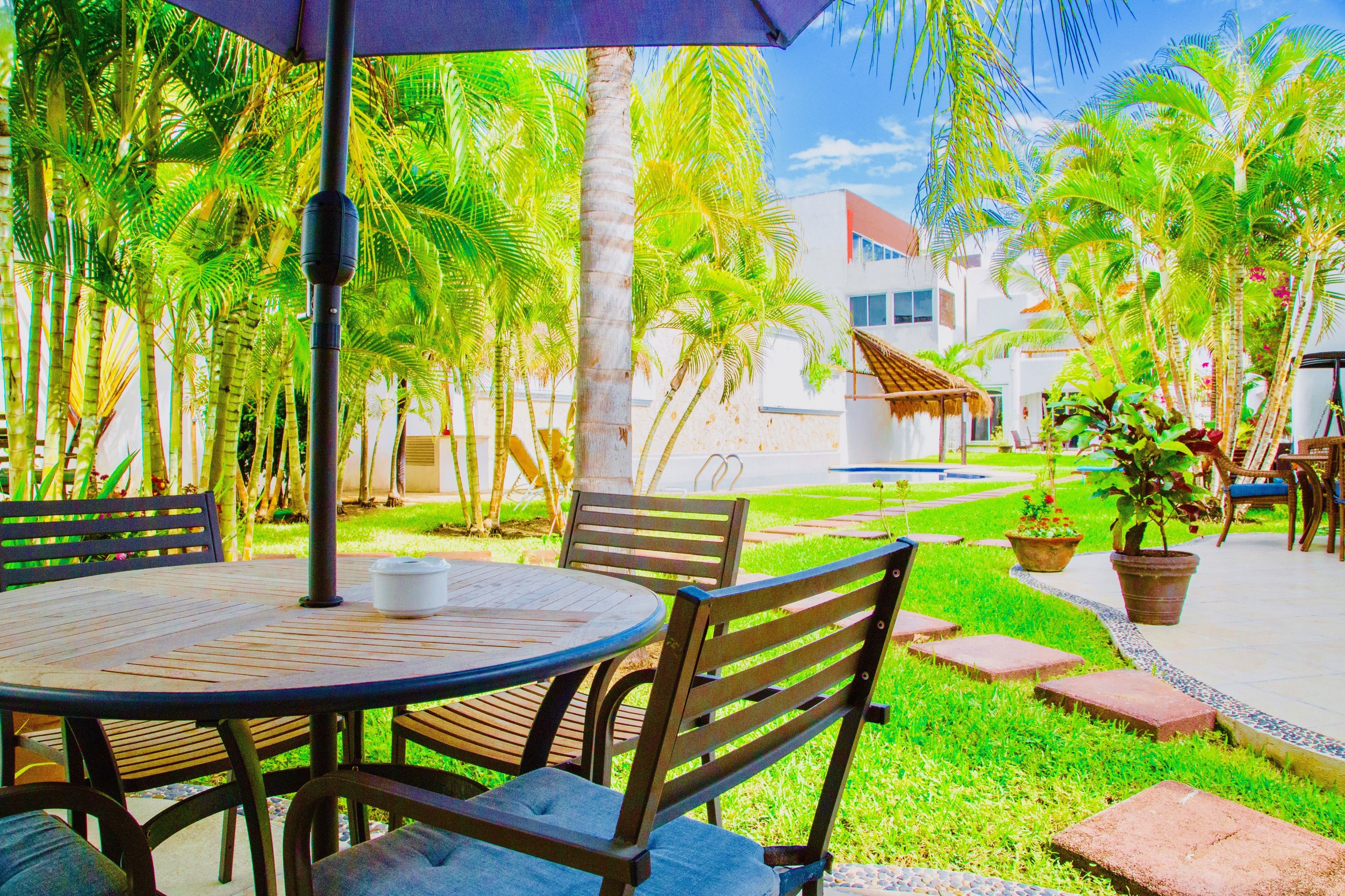A round wooden table with chairs, a thatched-roof structure, and a palm tree-lined pathway.