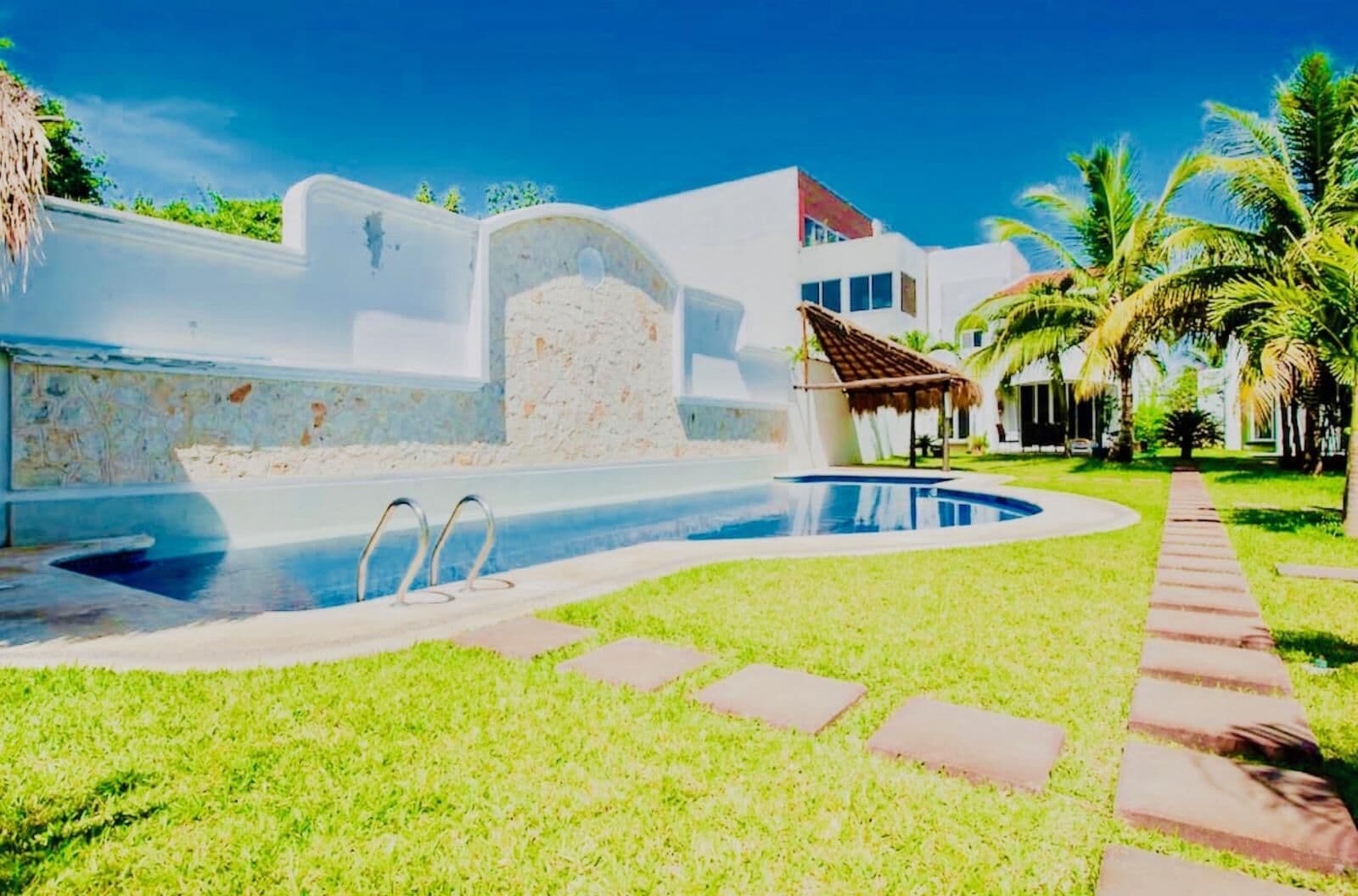 A swimming pool with a stone wall, a thatched-roof structure, and palm trees.