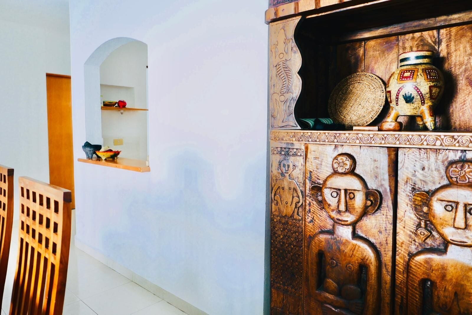 A room with a wooden cabinet featuring carved faces, a small shelf with decorative items, and a window with a wooden frame.
