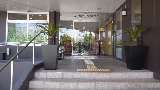 Entrada de hotel con una rampa que lleva a la puerta, plantas en macetas y fachada de vidrio transparente.