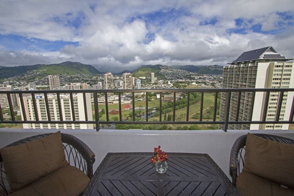 A balcony with wicker chairs and a table, overlooking a cityscape with mountains and buildings.