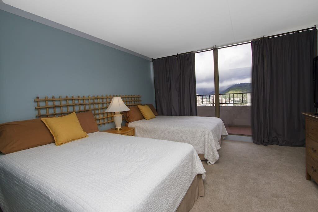 A hotel room with two beds, a wooden headboard, a nightstand with a lamp, and a view of the city through large windows.