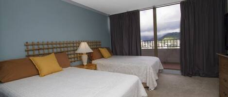 A hotel room with two beds, a wooden headboard, a nightstand with a lamp, and a view of the city through large windows.