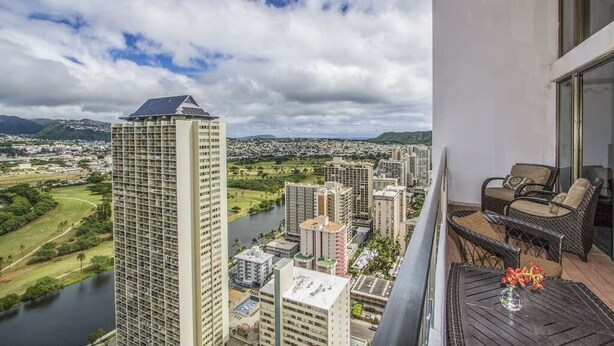 A balcony with wicker furniture and a vase of flowers, overlooking a cityscape with a river and a tall building. - Royal Hawaiian Hospitality (Honolulu)