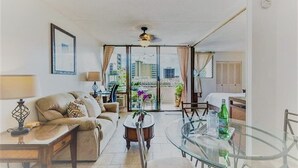 A modern living room with a sofa, coffee table, and glass dining table. There is a balcony with plants and a view of buildings. - Royal Hawaiian Hospitality (Honolulu)